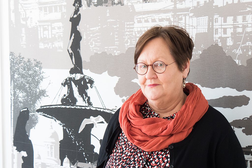 Leena Leppänen. Taustalla kangas jossa kaupunkinäkymä Helsingin Kauppatorilta.