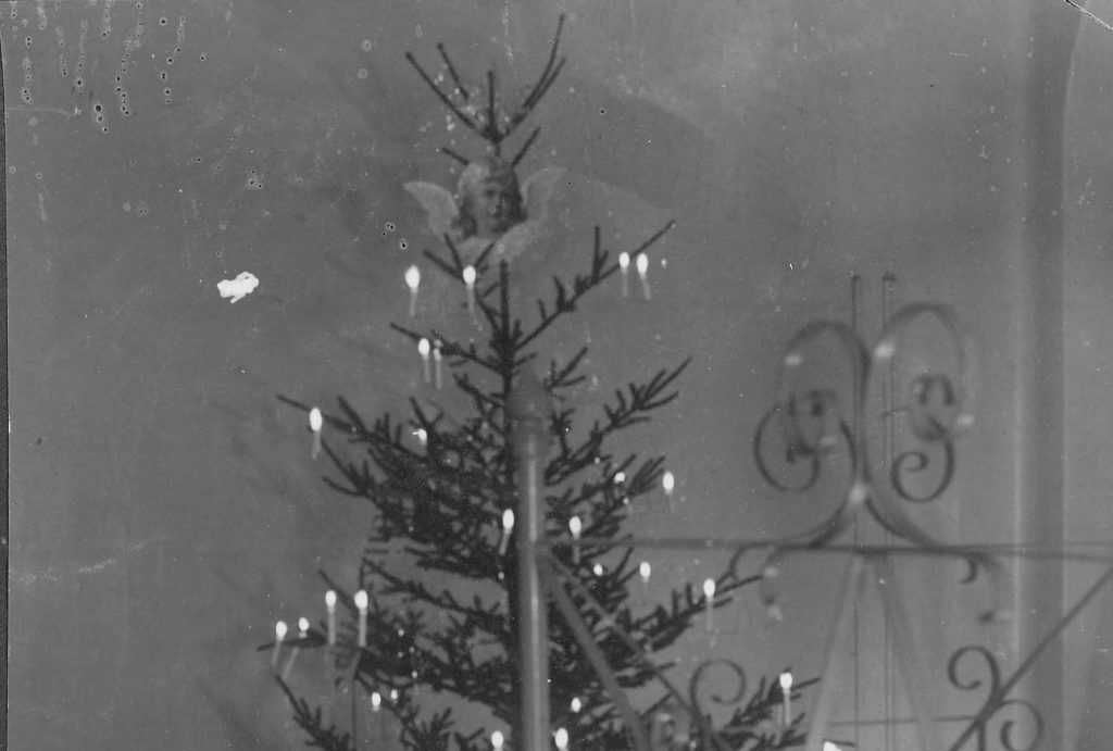 Mustavalkoisessa valokuvassa on joulukuusi, jonka latvassa on enkelikoriste ja oksilla sytytettyjä kynttilöitä. 