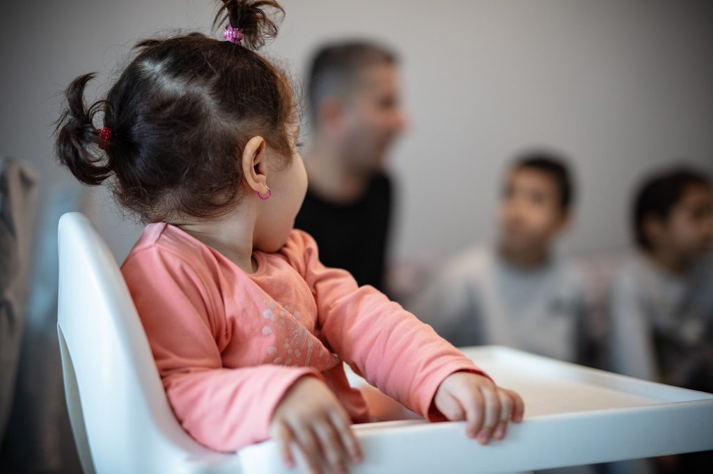 Pieni tyttö istuu syöttötuolissa, taustalla isä ja kaksi poikaa.