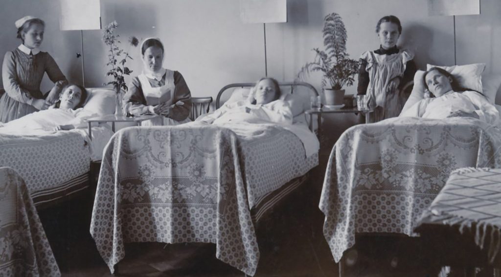 Kolme potilasta vuoteissaan, kahden vierellä on hoitaja ja yhden vierellä ehkä vierailija.