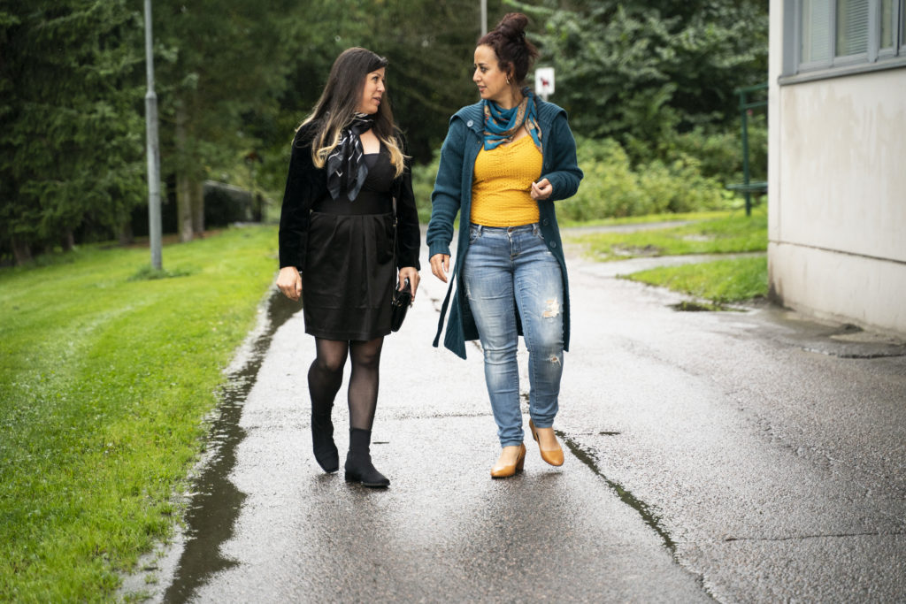 Kaksi naista kävelevät sateesta märällä asfalttitiellä, toistensa kanssa keskustellen.