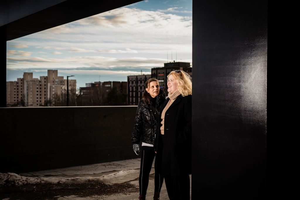 Kaksi naista seisoo katoksen alla, taustalla näkyy kaupunkimaisemaa. Amigo-mentorointi yhdistää heitä.