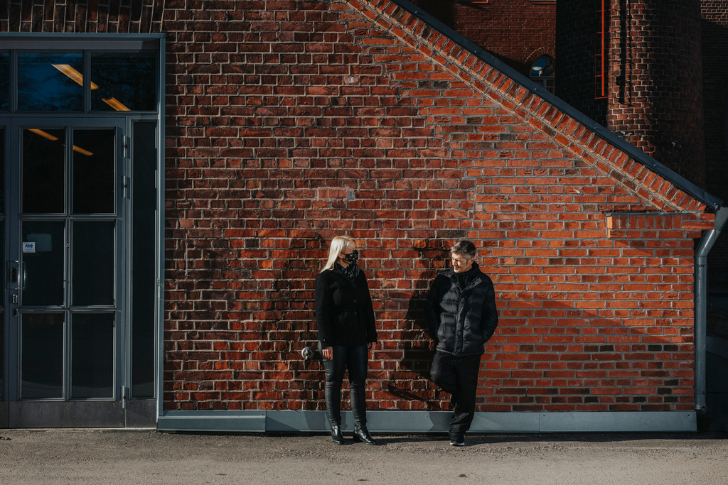 Kaksi henkilöä seisoo punatiilisen seinän edustalla.
