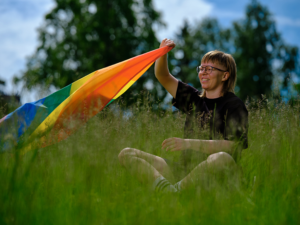 Henkilö istuu pellolla ja liehuttaa sateenkaarilippua.