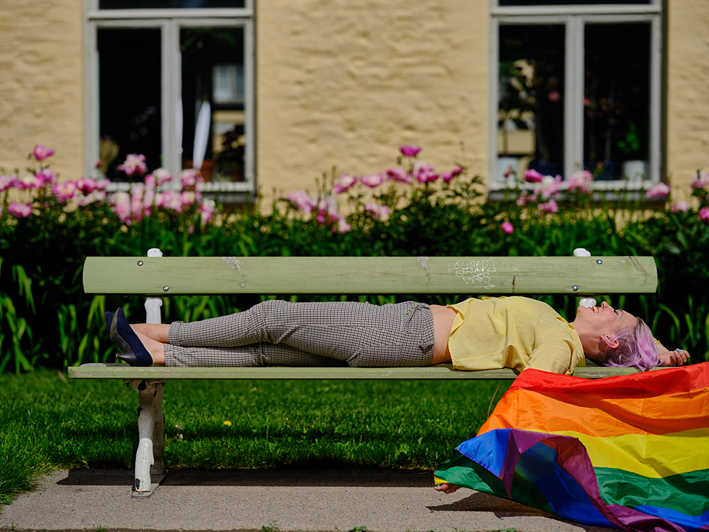 Ihminen makaa puistonpenkillä ja pitää kädessään sateenkaarilippua.