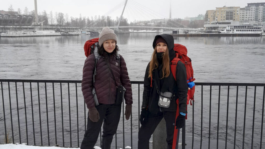 Kaksi nuorta henkilöä seisoo sillan kaiteen edessä, taustalla vesistö ja kaupunkimaisema. Tukialus Tampere on heidän työpaikkansa.