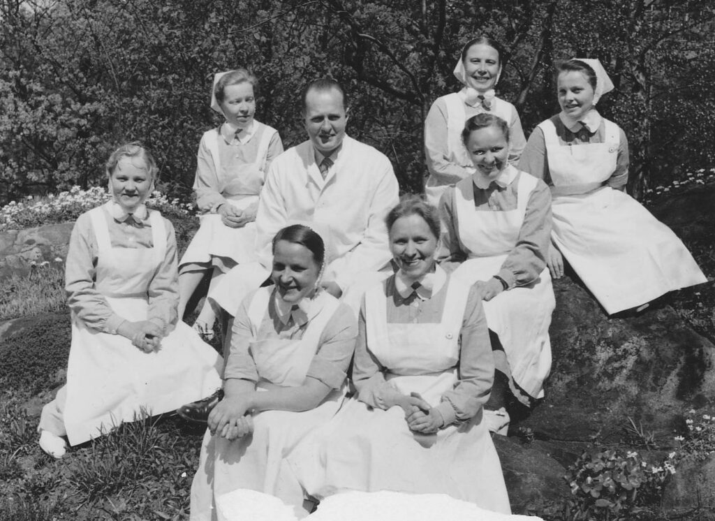 Ryhmä naisia istuu mieslääkärin ympärillä. Naiset ovat diakonissa-sairaanhoitajia.