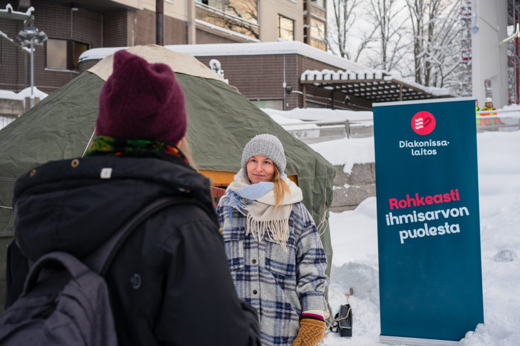 Kaksi naista keskustelee. Taustalla luminen maisema, jurtta ja sininen roll up, jossa on Diakonissalaitoksen logo ja teksti "Rohkeasti ihmisarvon puolesta."