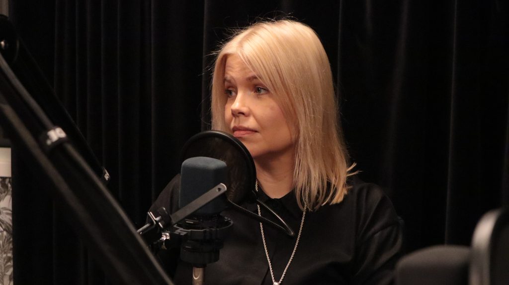 Vaaleahiuksinen nainen istuu mikrofonin takana.