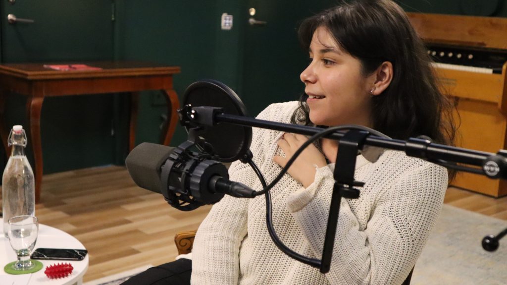 Nuori tummahiuksinen nainen hymyilee mikrofonin takana podcast-studiossa.