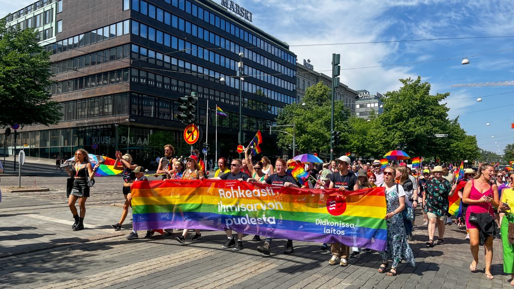 Pride-kulkue. Joukko ihmisiä kävelee kadulla kantaen sateenkaaren väristä banneria, jossa on teksti "Rohkeasti ihmisarvon puolesta".
