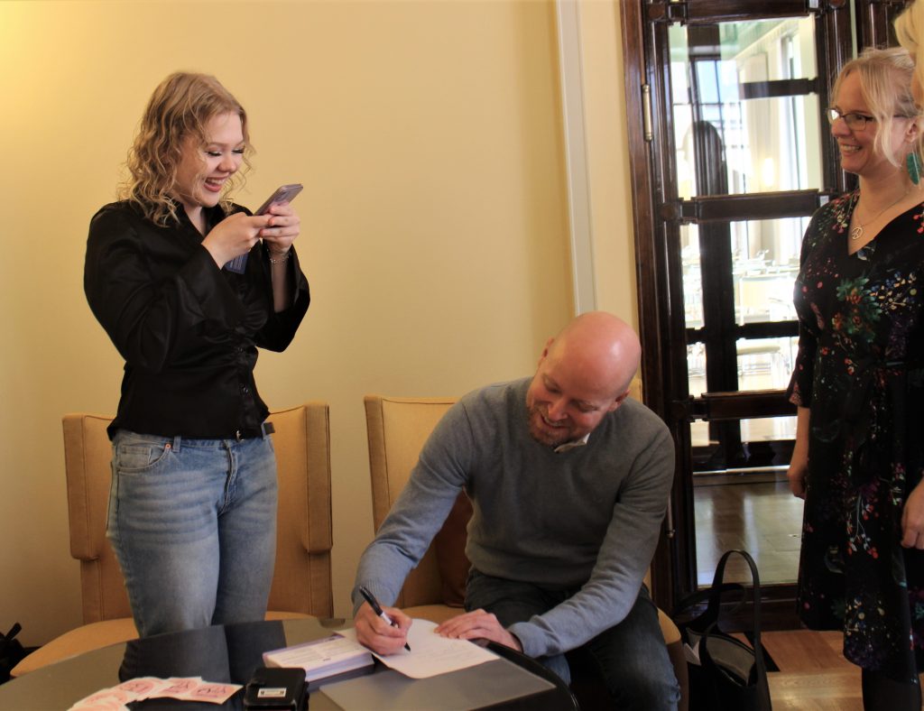 Jussi Saramo allekirjoittaa pöydän ääressä lakialoitetta ja Mila ottaa siitä kännykällä kuvaa hymyillen.