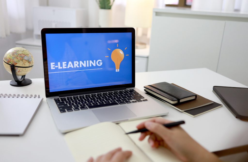 A laptop with a text e-learning on the screen and hands of a person taking notes.