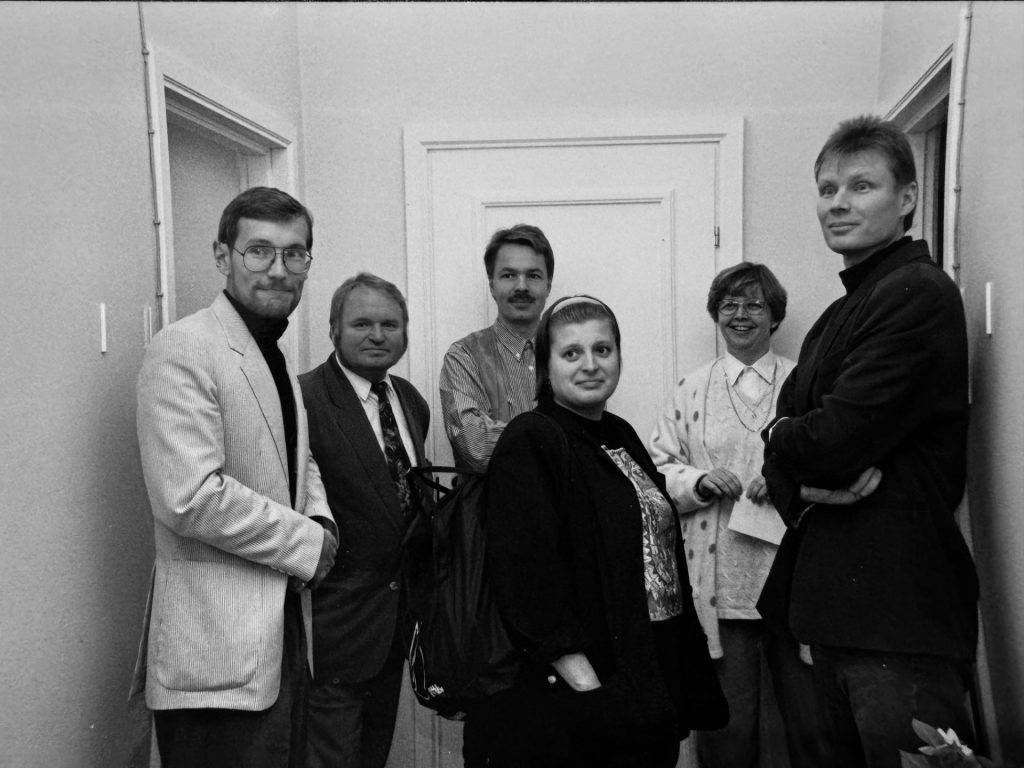 Mustavalkoinen kuva, jossa kuusi henkilöä seisoo käytävällä ja katsoo kameraan hymyillen.