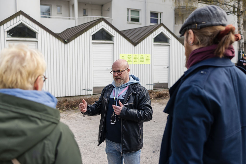 Mustaan nahkatakkiin pukeutunut mies keskustelee kahden ihmisen kanssa.