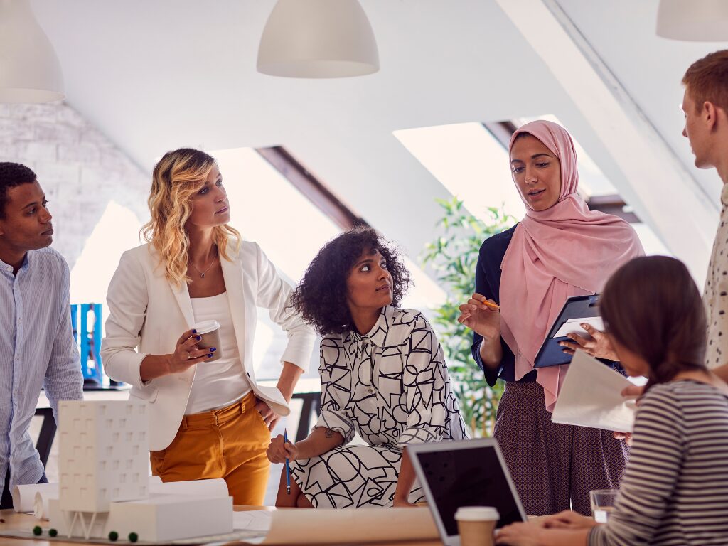 Five people standing and one sitting in an office setting, listening to a woman wearing a hijab.