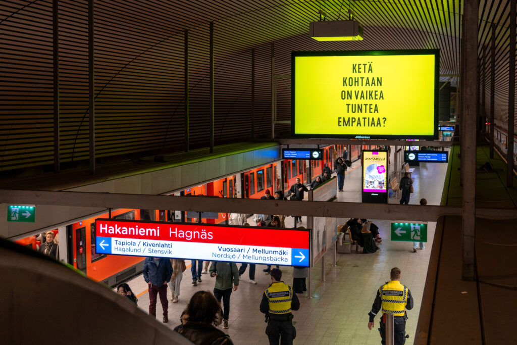 Metrolaituri, jolla kulkee ihmisiä. Laiturin yläpuolella olevassa digitaalisessa mainostaulussa on keltaisella pohjalla teksti: "Ketä kohtaan on vaikea tuntea empatiaa?".