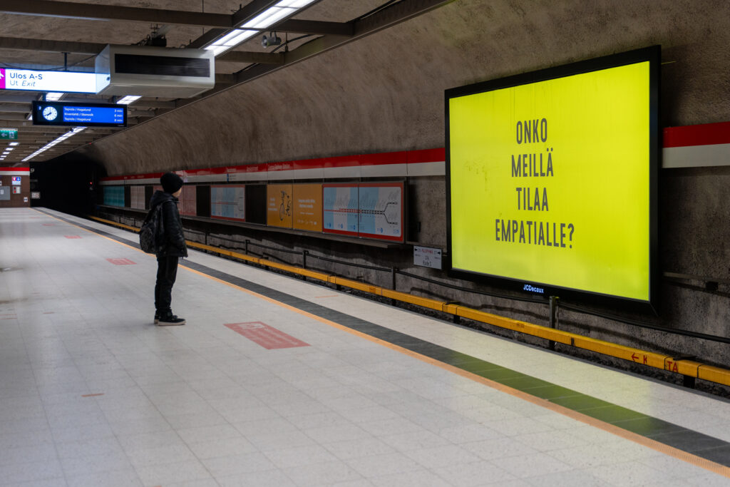 Poika seisoo yksin metrolaiturilla ja katsoo seinällä olevaa digitaalista mainostaulua, jossa keltaisella pohjalla lukee teksti: "Onko meillä tilaa empatialle?".
