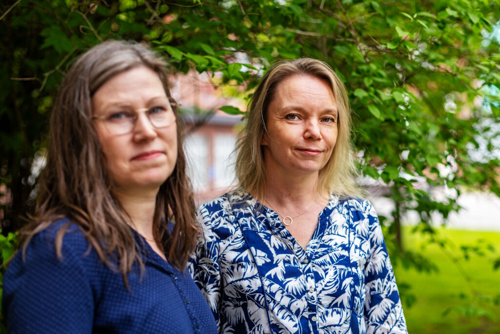 Kaksi naista seisoo rinnakkain ja katsoo vakavina kameraan. Taustalla punatiilinen rakennus ja vihreää kasvustoa.
