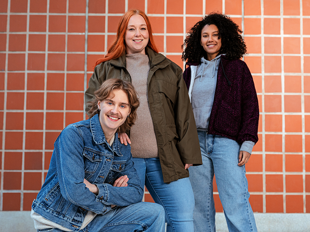 kolme nuorta aikuista seisoo punaisen tiiliseinän edessä hymyillen.