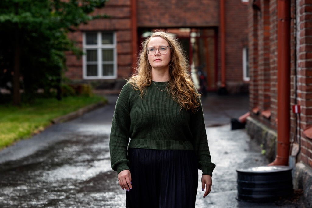 Vihreään villapaitaan pukeutunut, ruskeahiuksinen ja silmälasipäinen nainen seisoo asfaltoidulla pihalla. Taustalla punatiilinen rakennus.
