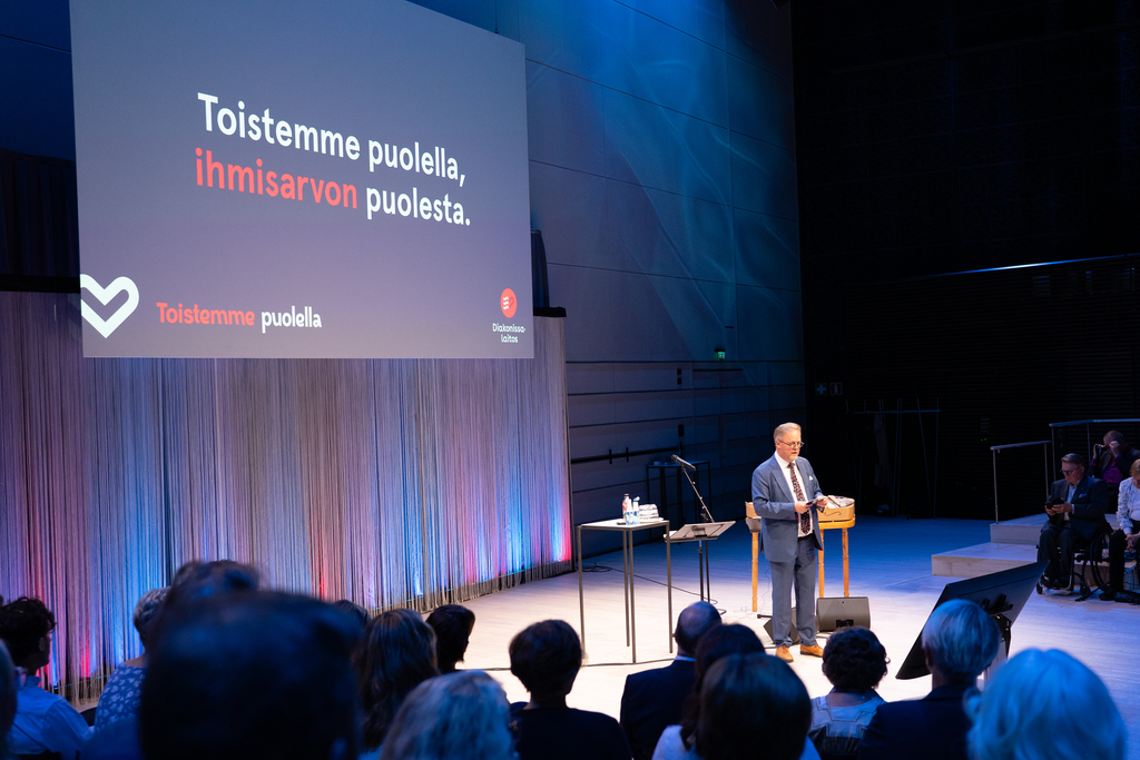 Puhuja salissa ja näytöllä teksti: Toistemme puolella, ihmisarvon puolesta