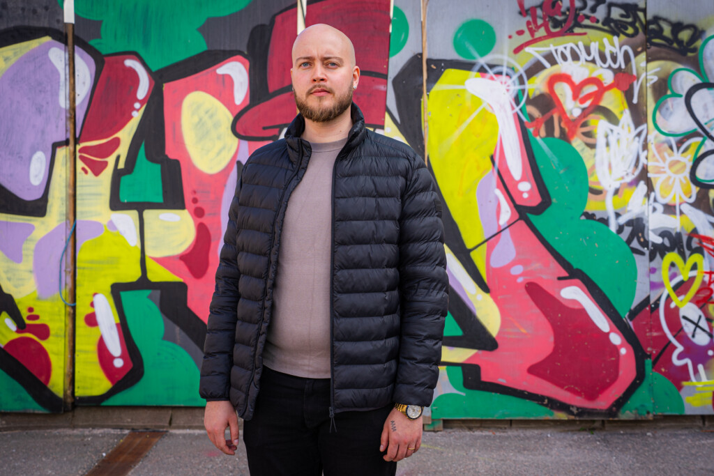 Mustiin housuihin, mustaan untuvatakkiin ja beigeen paitaan pukeutunut mies katsoo vakavana kameraan. Taustalla graffitiseinä.