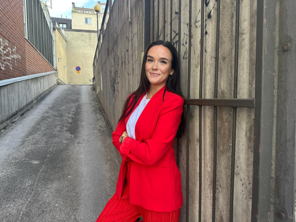 Punaiseen jakkupukuun pukeutunut nainen nojaa seinään ja katsoo hymyillen kameraan.