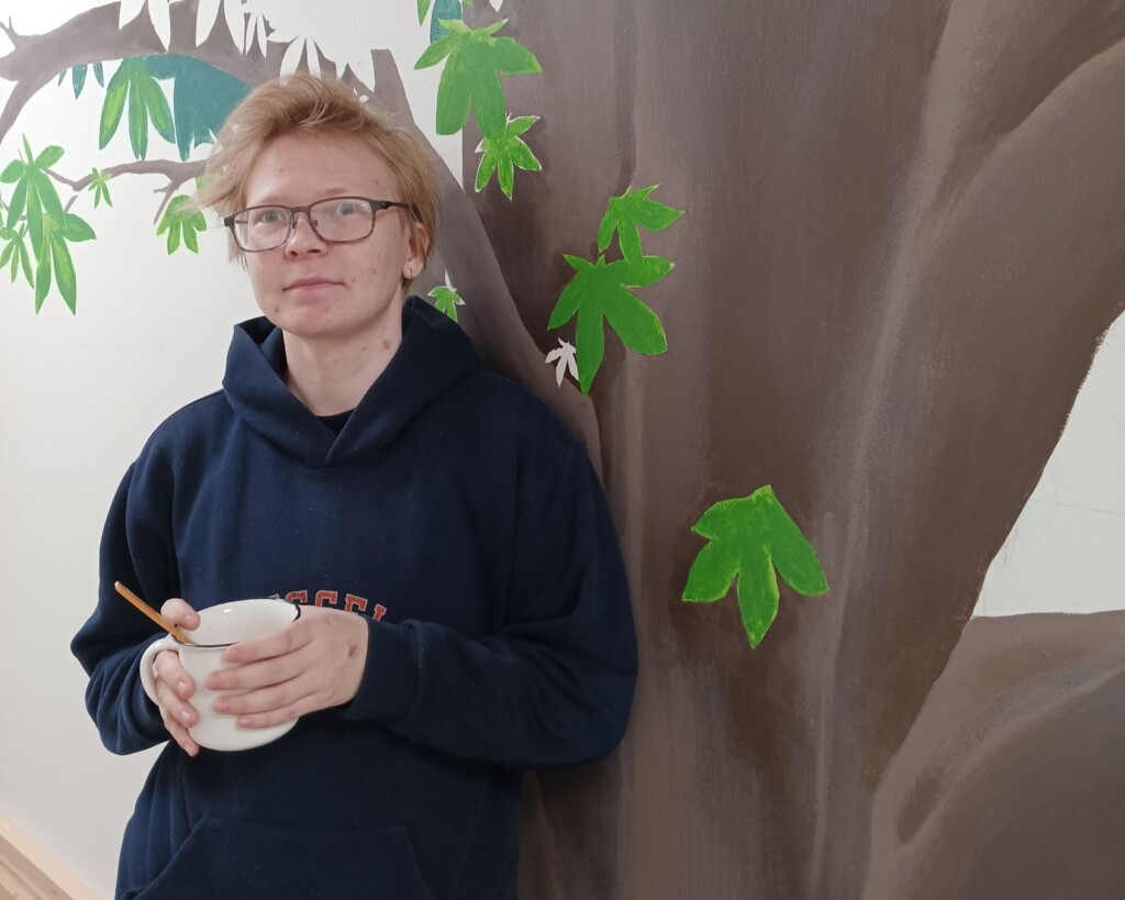 Silmälasipäinen, lyhythiuksinen ja tummaan huppariin pukeutunut nuori seisoo puuta esittävän muraalin edessä ja pitää käsissään kahvikuppia.