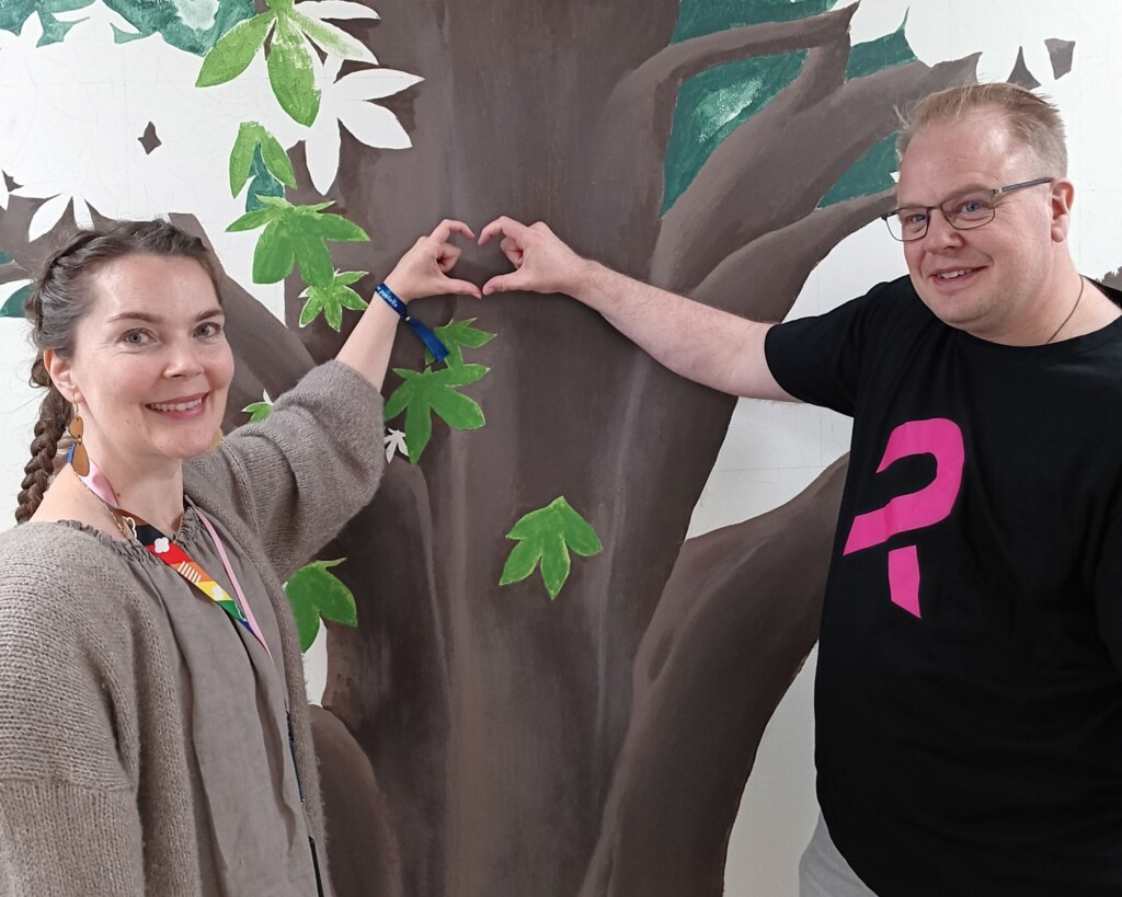 Nainen ja mies katsovat kameraan hymyillen ja muodostavat käsillään sydämen.