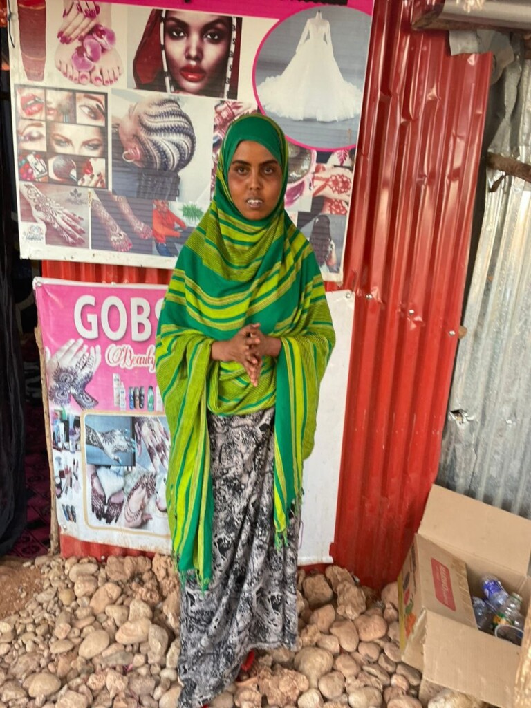 A young woman in a big green scarf and a dress stands in front of  a wall of corrugated iron covered with beaty care posters.