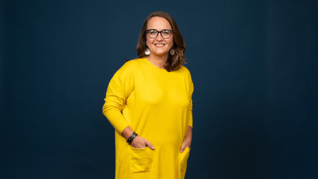 Keltaiseen mekkoon pukeutunut nainen katsoo hymyillen kameraan.