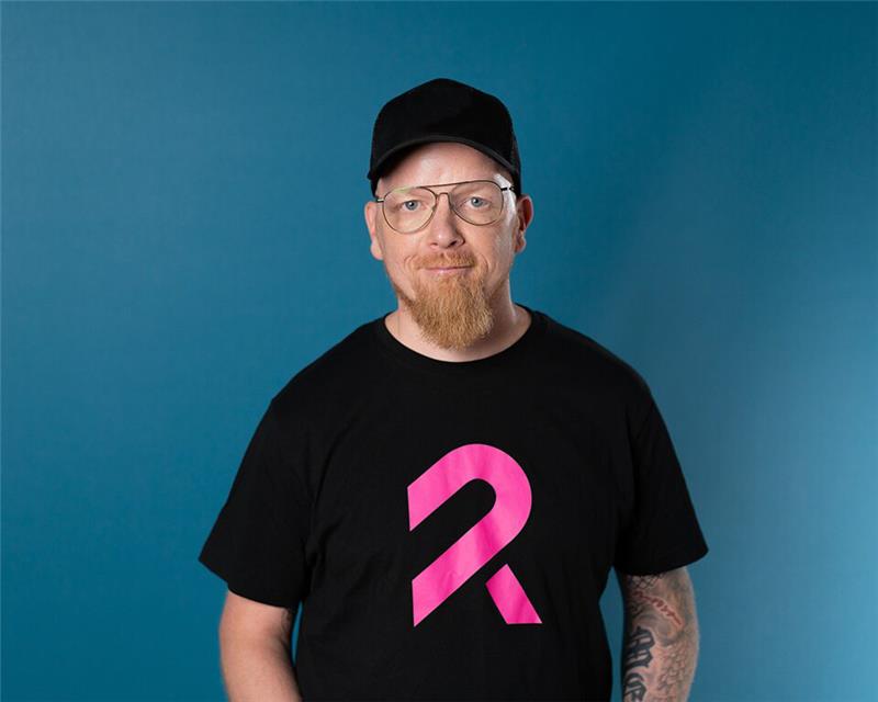 Mustaan t-paitaan ja lippalakkiin pukeutunut silmälasipäinen mies katsoo hymyillen kameraan.