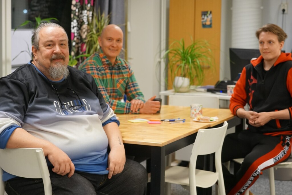 Kolme hyväntuulista miestä istuu pöydän ääressä ja katsoo kameraan.