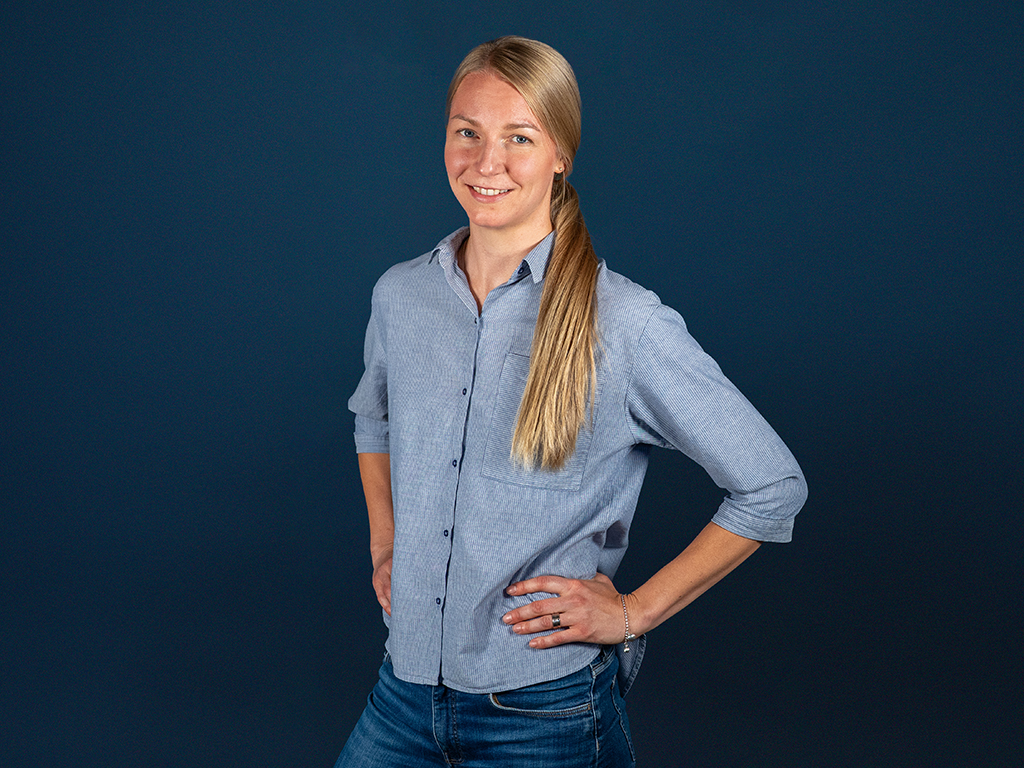 Nainen seisoo hymyillen ja kädet lanteille sinistä seinää vasten.