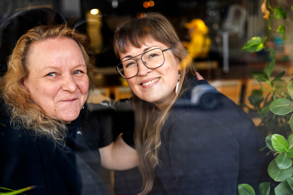 Riitta ja Virpi istuvat vierekkäin ja hymyilevät.