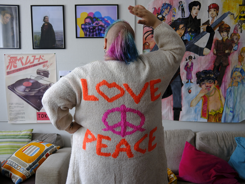 Nuori aikuinen seisoo selin päällään neuletakki, jossa lukee "Love and Peace".