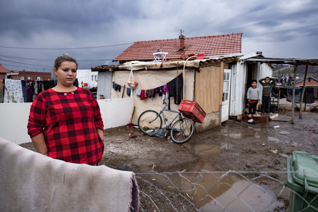 Nainen seisoo mutaisen pihan reunalla, jonka toisella puolella olevan pienen talon edessä seisoo kaksi lasta.
