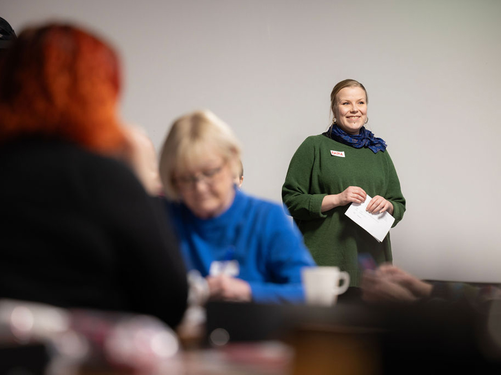 Nainen seisoo hymyillen ja paperi kädessään ihmisten ympärillä.